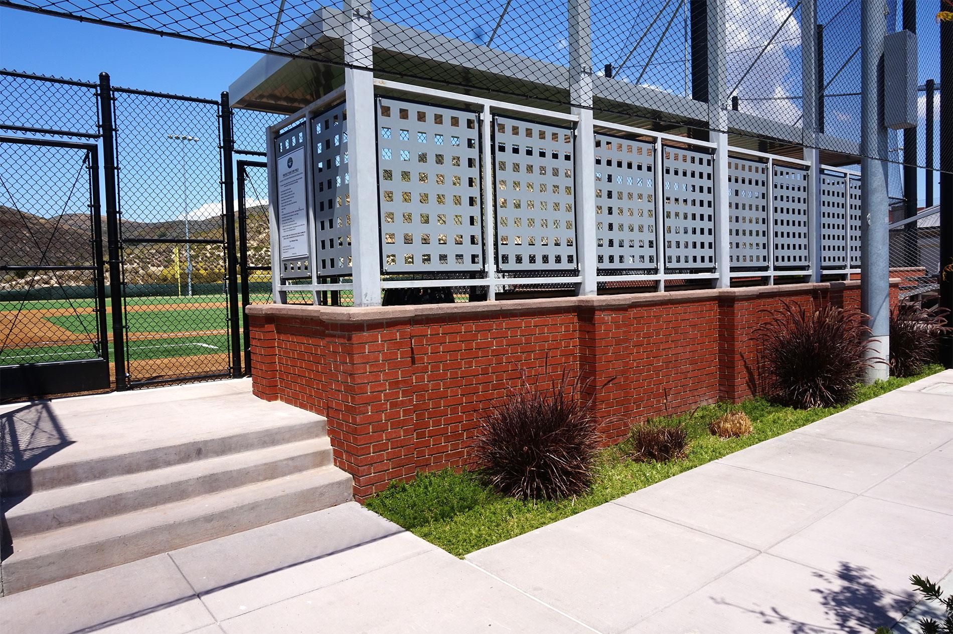 Red brick dugout wall topped with a white perforated steel screen, concrete steps and sidewalk alongside a baseball diamond