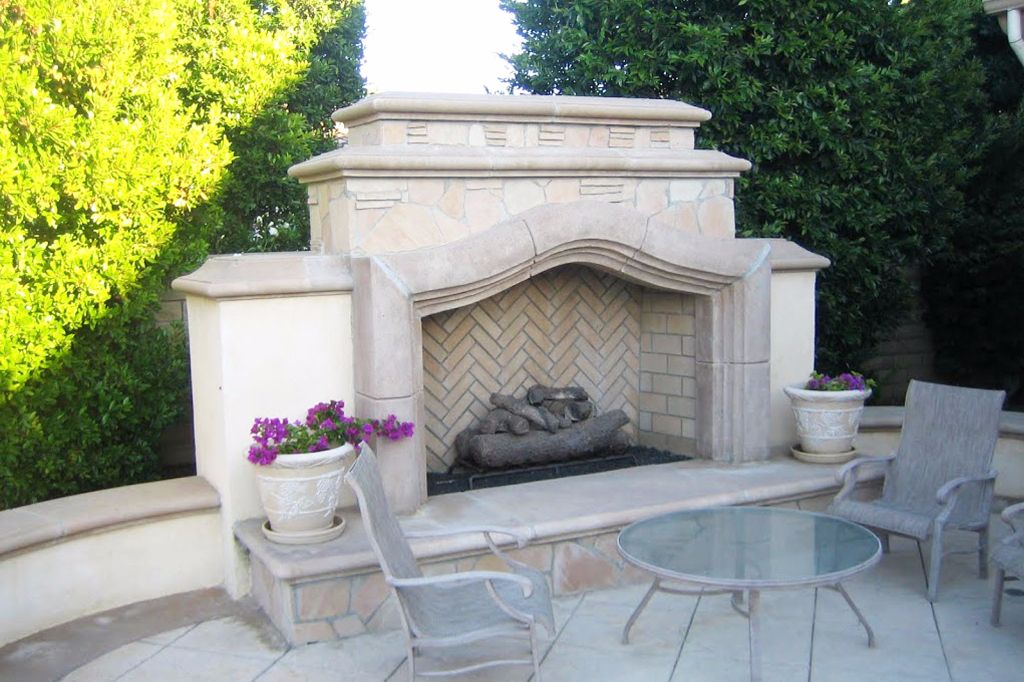 Cast stone outdoor fireplace with a scrolled mantel and herringbone brick firebox, flanked by a curved seat wall on a flagstone patio