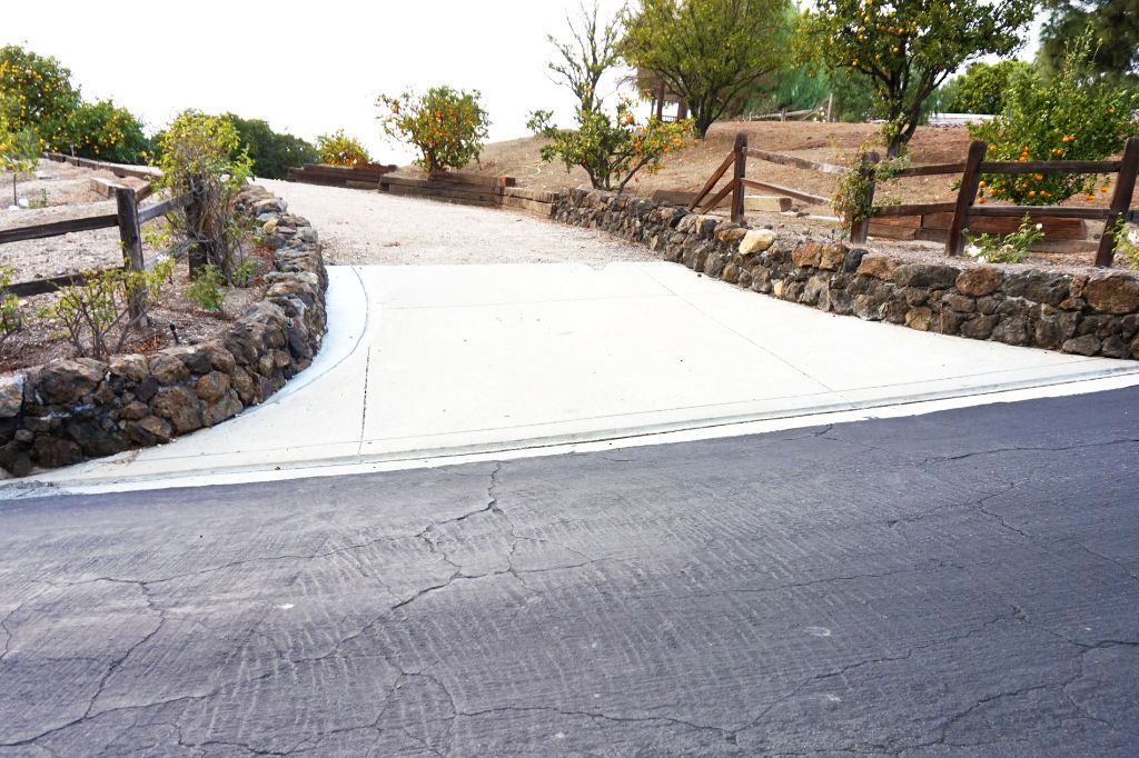 Concrete driveway curving down to the street between low stone retaining walls and a ranch fence