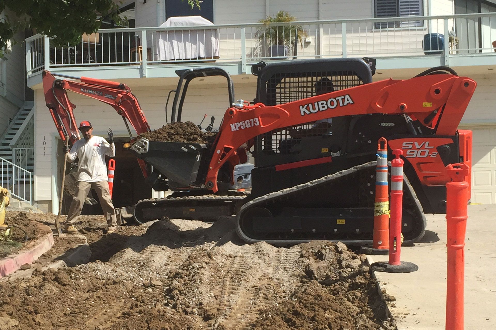 Mini excavator and track loader stripping a front yard, traffic cones set out along the street