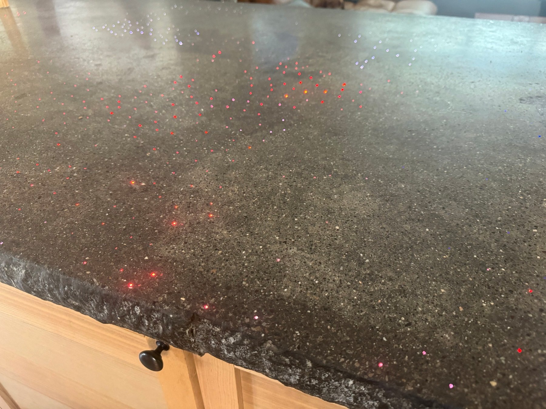 Fiber-optic concrete countertop in daylight, lit points scattered across the polished gray slab above a chiseled rock-face edge and an oak cabinet