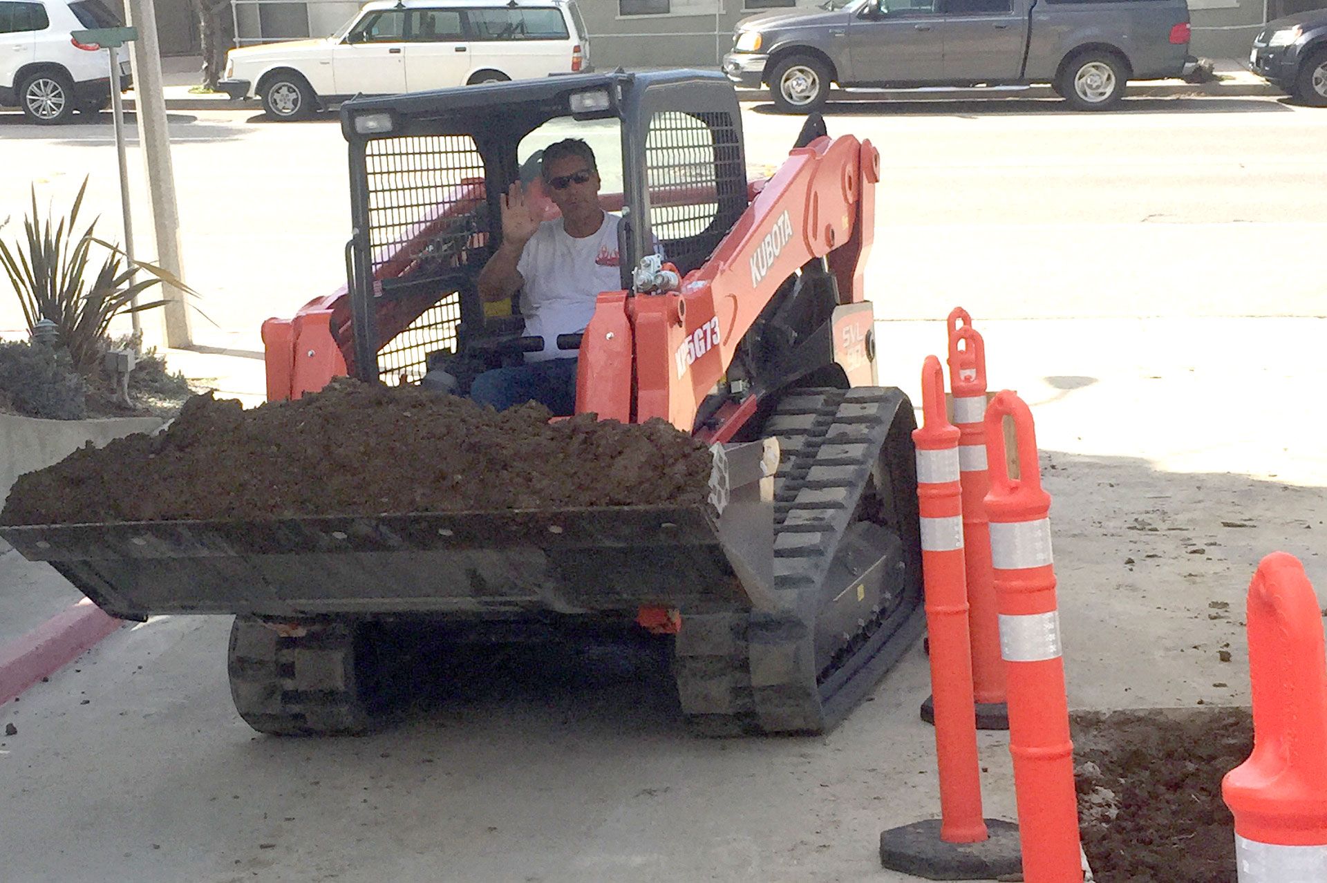 Operator moving a full bucket of excavated soil past a coned-off kerbside work area