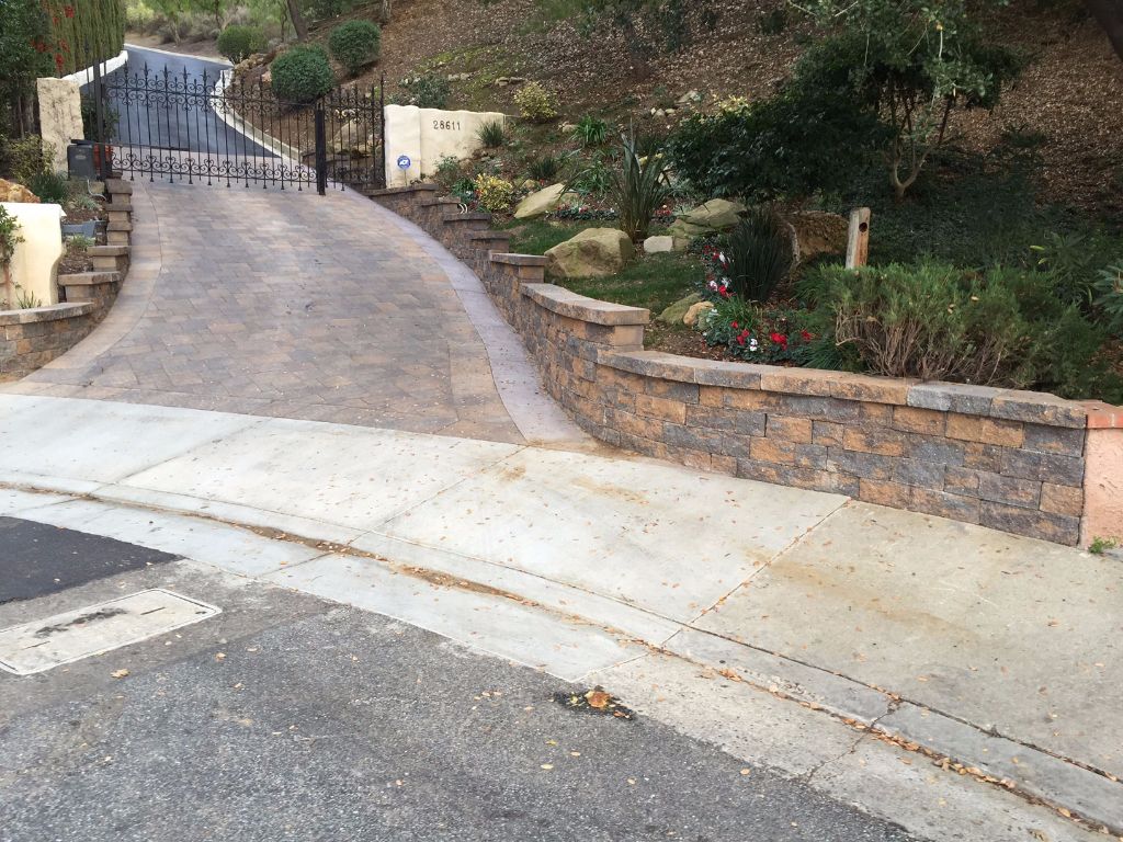 Paver driveway rising to an iron gate between block retaining walls with a cut stone cap and a planted slope behind
