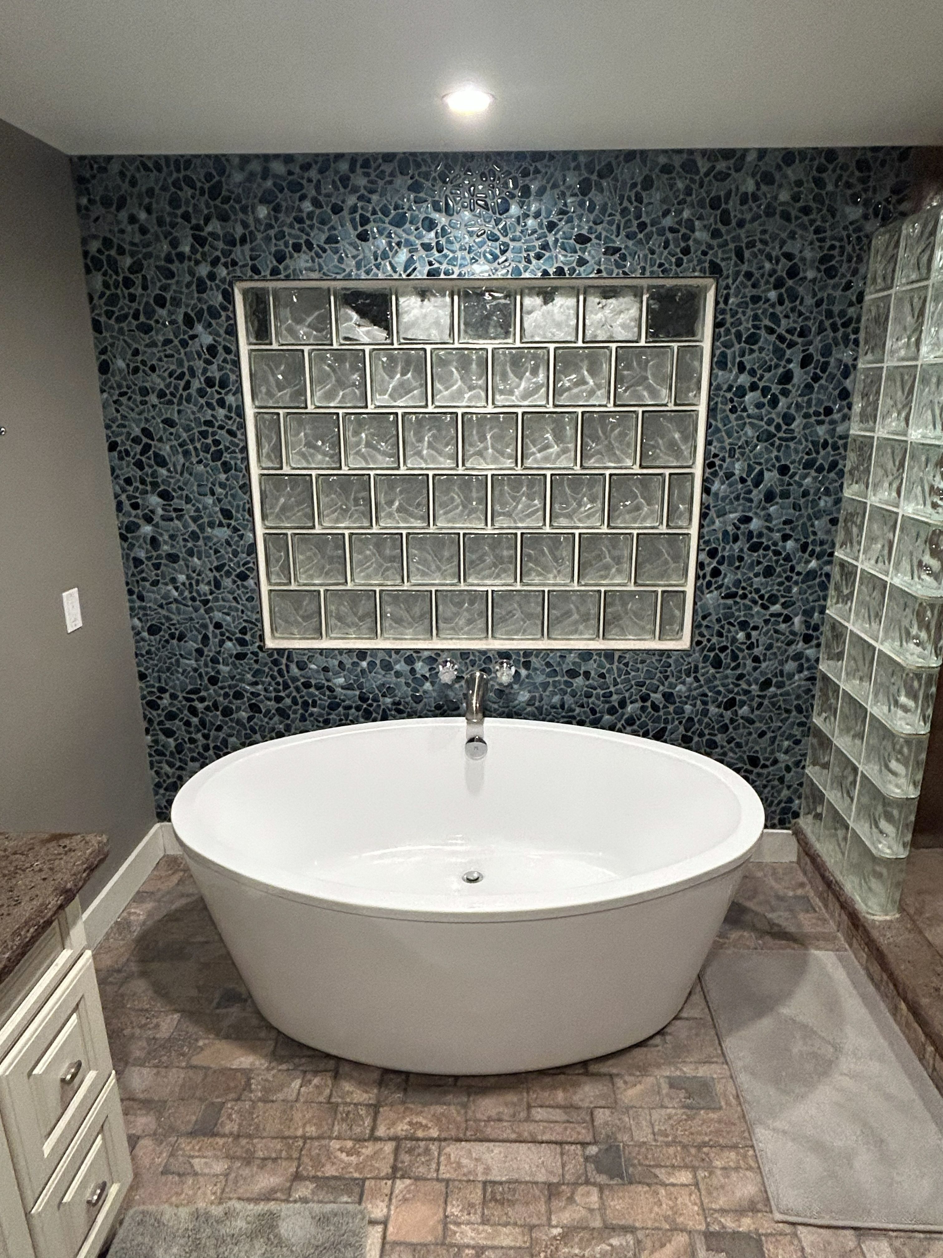 River rock feature wall and glass block window behind a freestanding bath, on stone tile