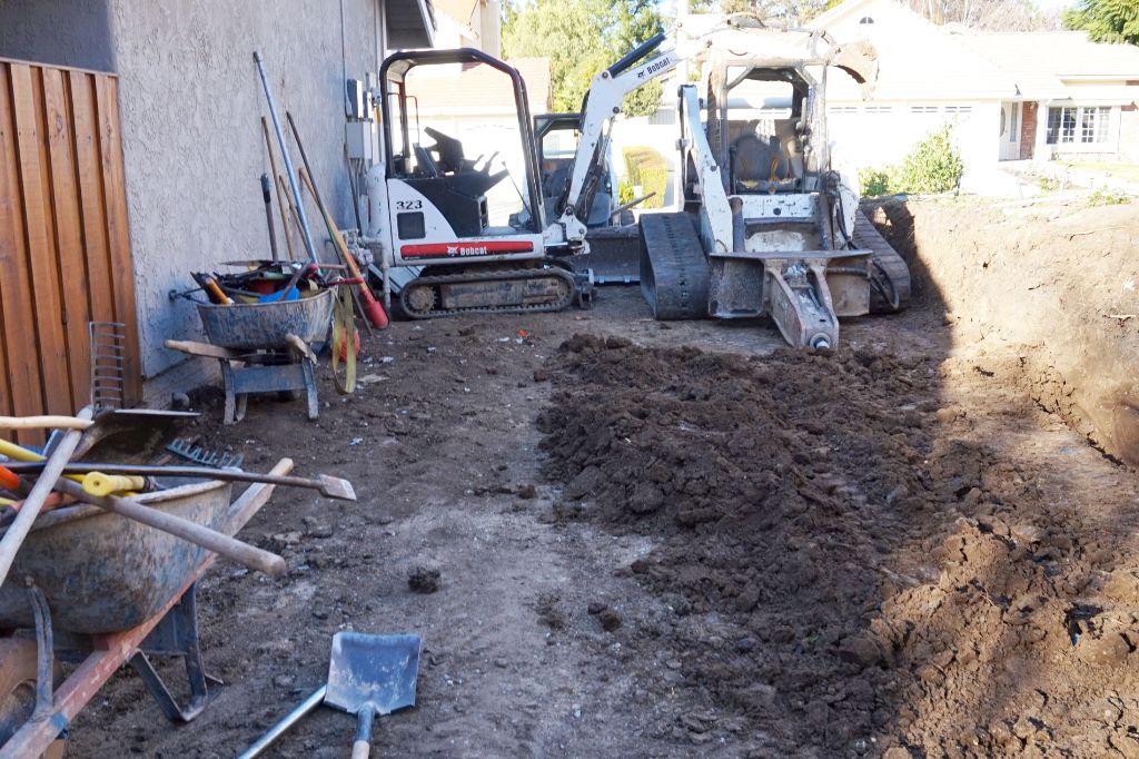 Skid-steer loader and hand tools working a narrow side yard between house and fence