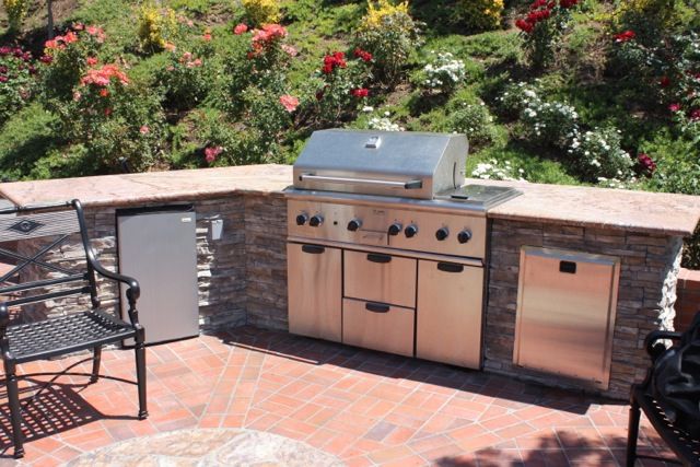Stacked stone barbecue island on a brick patio with a rose garden and climbing roses behind