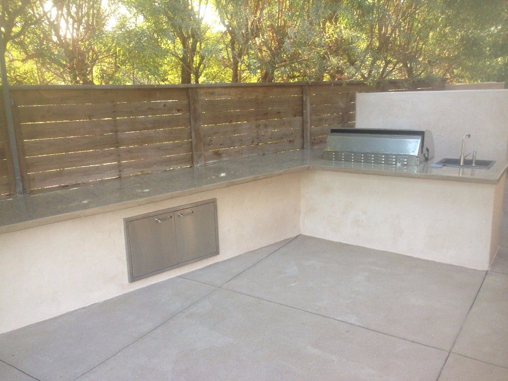 L-shaped stucco outdoor kitchen run along a slatted timber fence, granite counter over stainless access doors