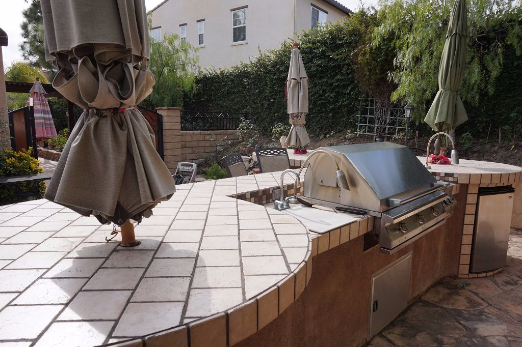Curved tiled outdoor kitchen counter with a built-in grill and refrigerator, laid out on a flagstone patio